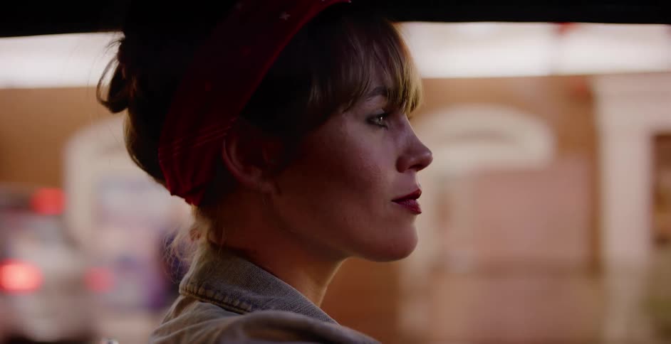 A pensive woman with a red bandana drives through the city at night, with blurred lights reflected in the car window