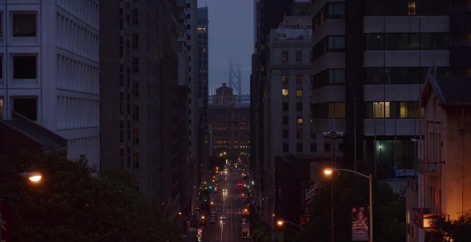 An atmospheric aerial view slowly pulls back from a quiet city street in San Francisco at dusk, with the Bay Bridge looming in the distance