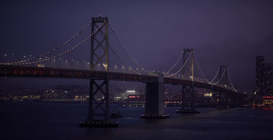 A cinematic reveal of the brightly illuminated San Francisco-Oakland Bay Bridge at night, with city lights twinkling in the background