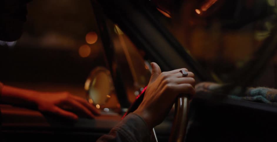 A driver grips the steering wheel of a vintage car, navigating through the vibrant, blurry city lights at night