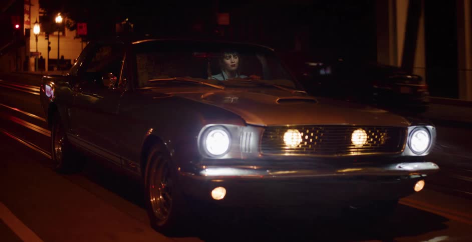 A woman cruises through the city at night in a classic vintage Ford Mustang with the headlights on