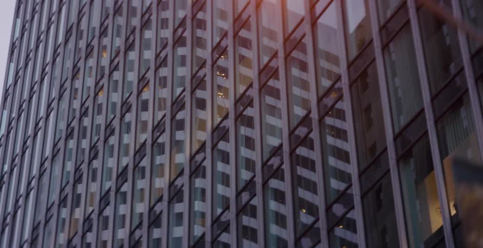 A slow pan across the reflective glass facade of a modern skyscraper at dusk reveals a streetlamp passing by