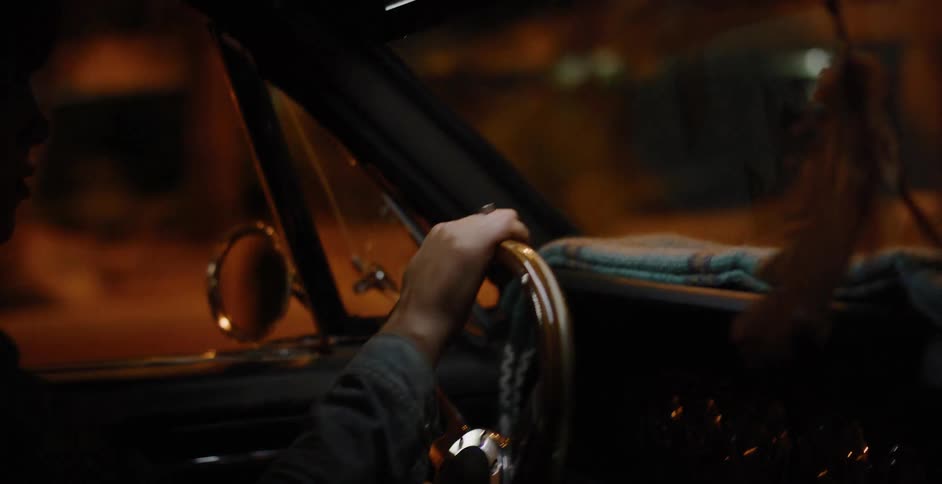 A driver's hand rests on the steering wheel of a classic car during a moody drive through the city at night