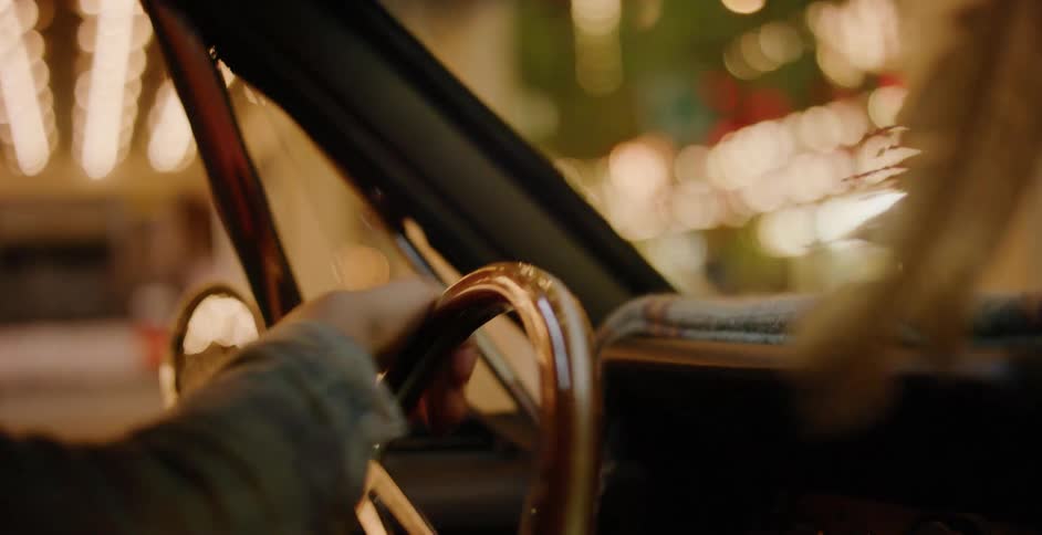 A close-up of a person's hands on the wooden steering wheel of a vintage car driving through a city with bokeh lights at night