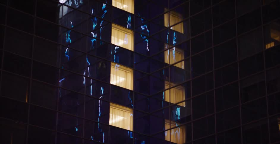 Warmly lit windows dot the reflective glass facade of a modern skyscraper as it captures the city's neon lights at night