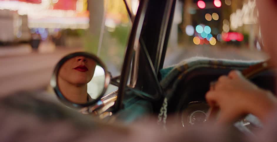 A woman's reflection with red lipstick is visible in the side-view mirror of a vintage convertible as she drives through a city at night