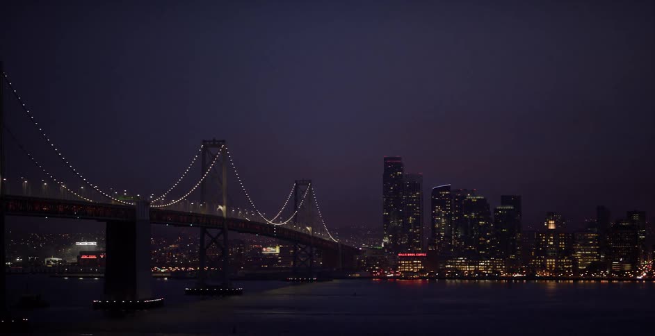 An aerial tracking shot reveals the beautifully lit San Francisco Bay Bridge and downtown skyline at night