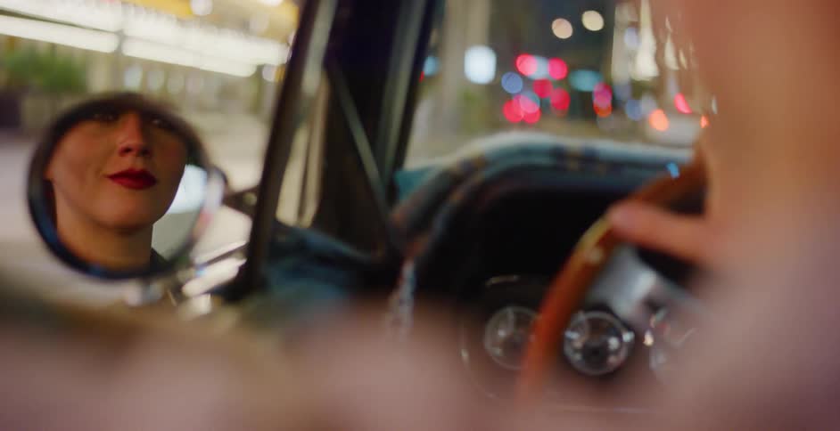 A woman's reflection with red lipstick is visible in a classic car's side-view mirror during a night drive through the city