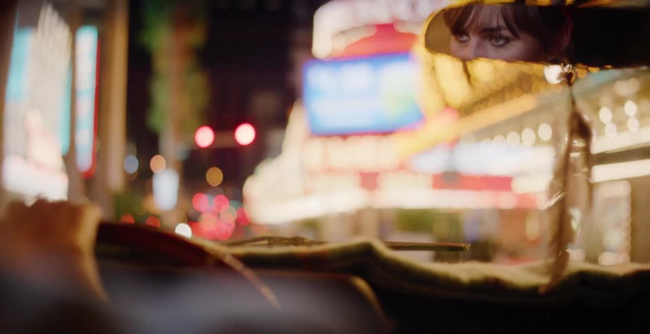 A woman's eyes are reflected in the rearview mirror of her car as she waits in traffic on a brightly lit city street at night