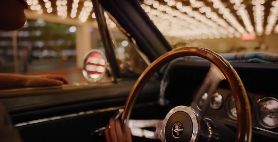 A person impatiently taps their fingers on the wooden steering wheel of a classic car while waiting under the bright bokeh lights of a city at night