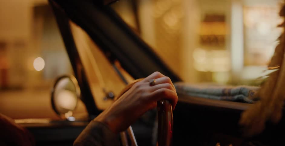 A close-up of a person's hand on the steering wheel while driving a vintage car through a city at night