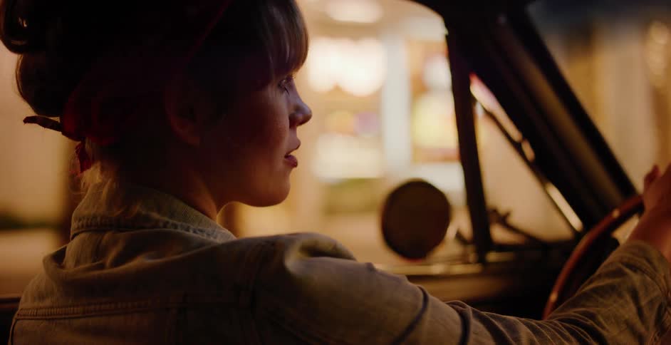 A young woman with a red bandana drives a classic car through the city at night, illuminated by the warm glow of streetlights