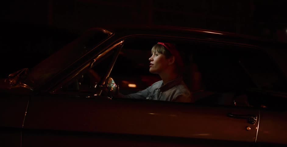 A stylish woman with a red bandana drives a classic American muscle car through the city at night