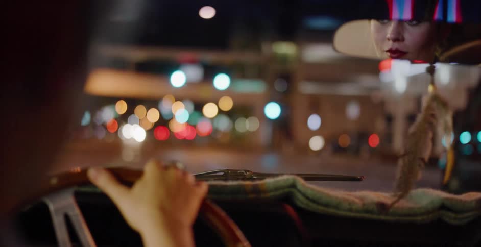 A woman's reflection is visible in the rearview mirror as she drives through a city at night with soft bokeh lights
