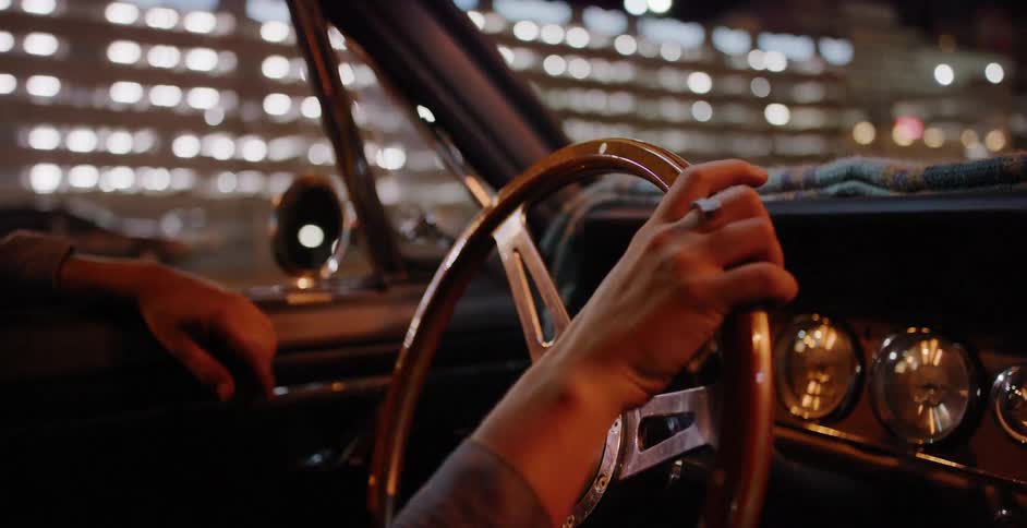 A driver grips the wooden steering wheel of a vintage convertible while cruising through the city at night with bokeh lights in the background