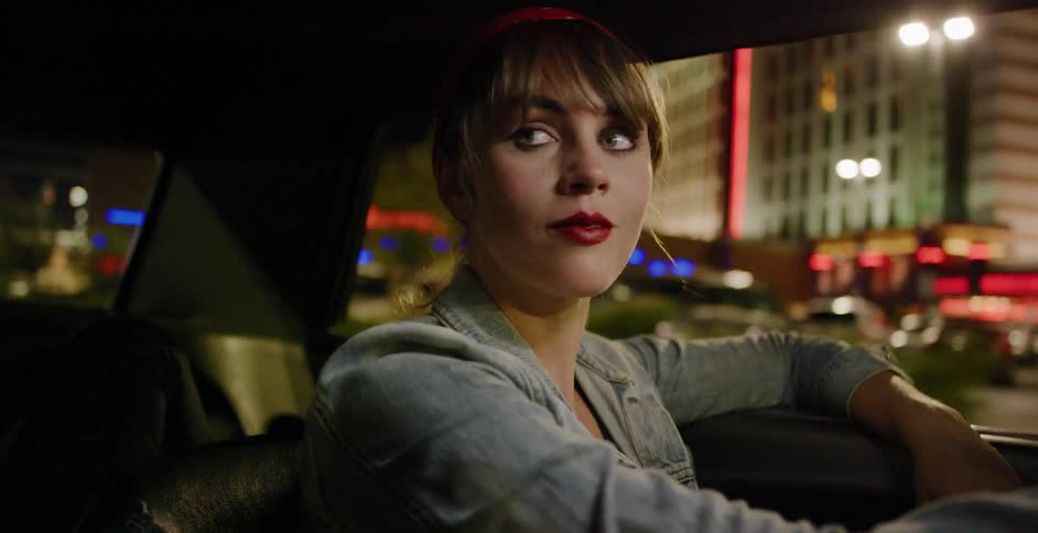 A fashionable woman in a denim jacket and red bandana takes in the city lights from the passenger seat of a moving car at night