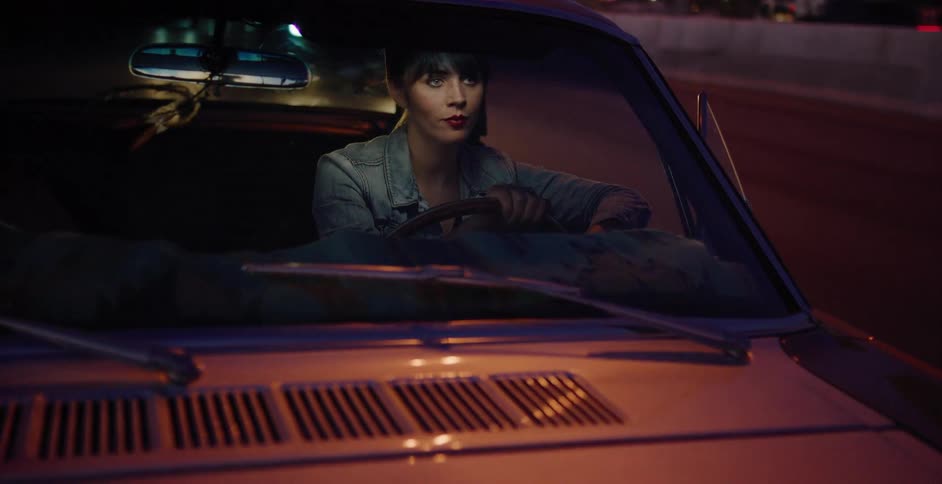 A stylish young woman drives a classic car along a highway at night as city lights reflect off the windshield