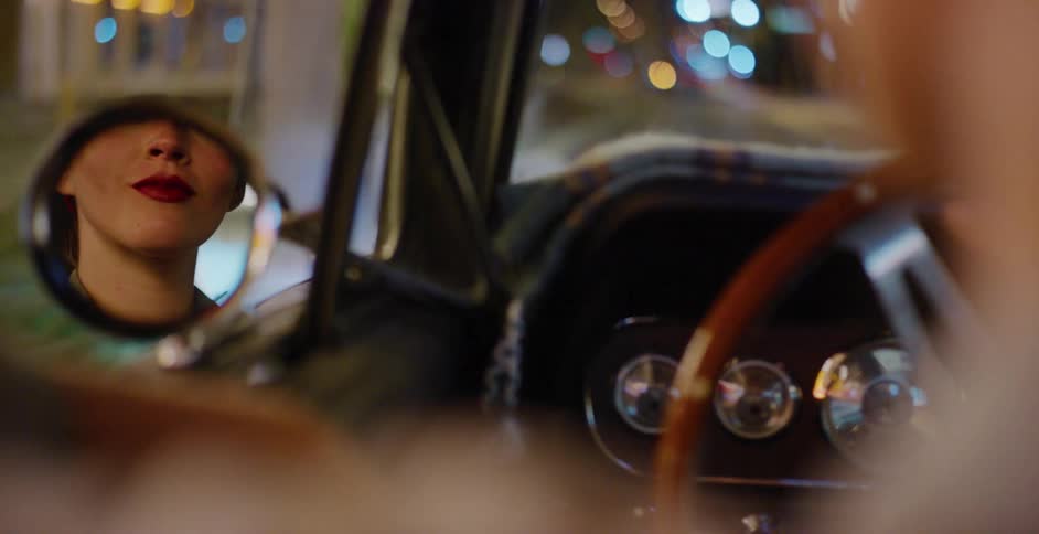 Close up on the reflection of a stylish woman in the side-view mirror of a classic car driving through a city at night