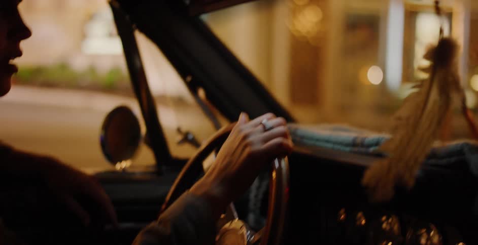 A woman's hands grip the steering wheel of a vintage car as she drives through the city at night