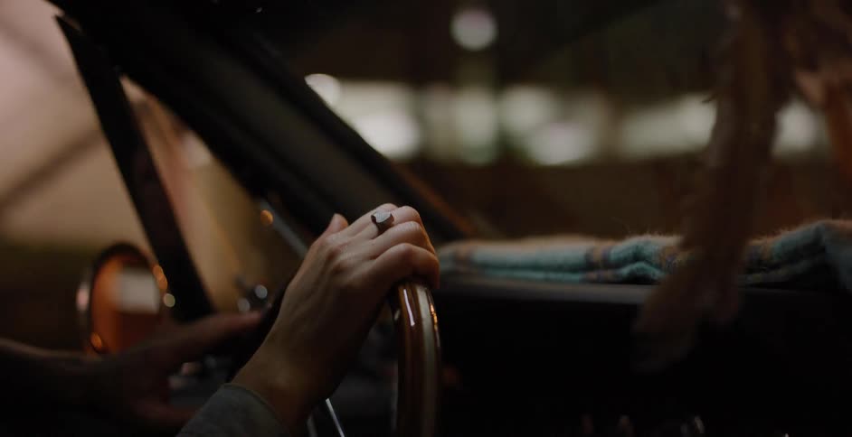 A close-up shot captures a driver's hand on the wooden steering wheel of a vintage car, navigating through the blurry city lights at night
