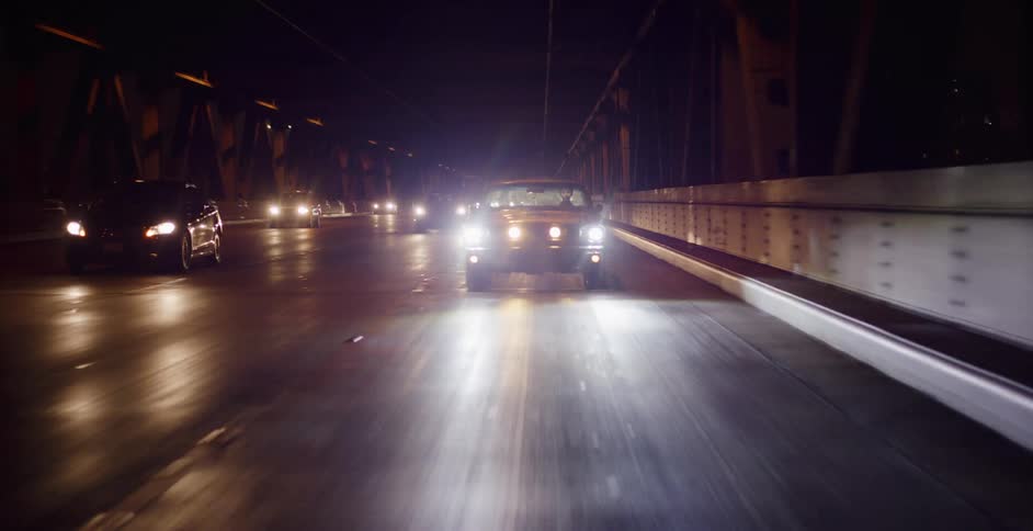 POV shot following a classic black convertible with bright headlights driving across a city bridge at night