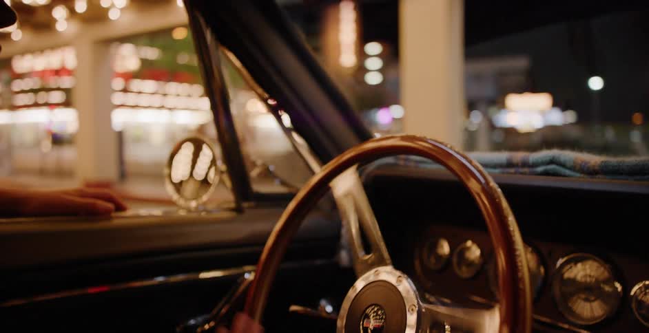 Interior shot of a person's hands on a vintage steering wheel while driving through a brightly lit city at night