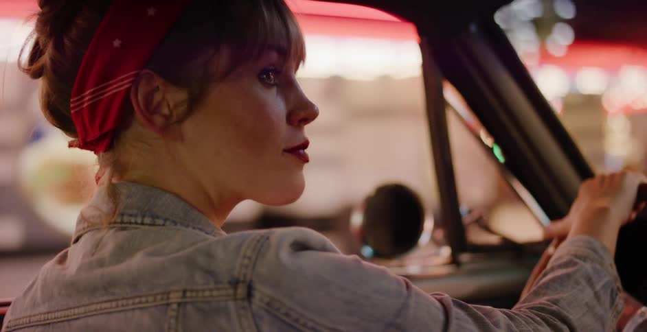 A stylish woman in a red bandana drives a vintage car past the glowing neon lights of the city at night