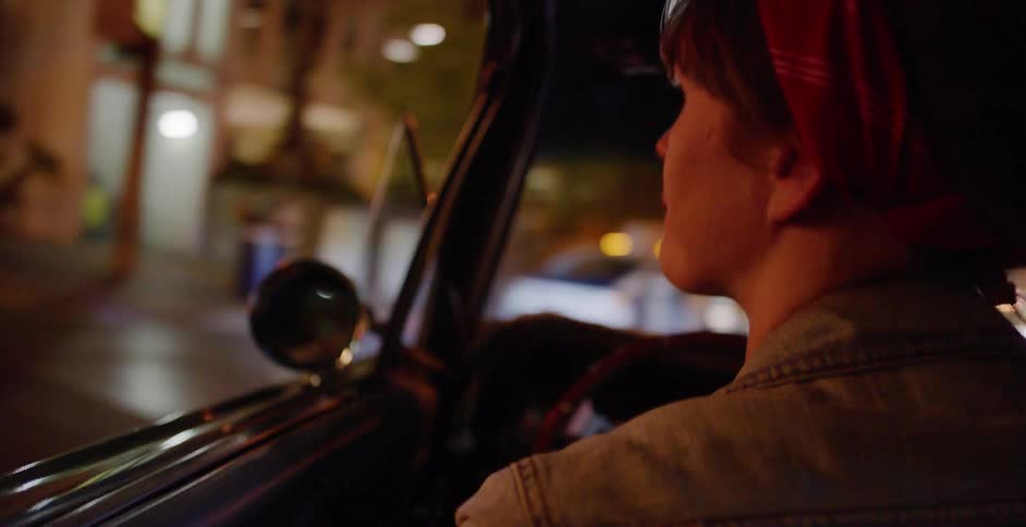 A woman in a denim jacket and red bandana drives a vintage convertible through the city at night as the background lights blur past