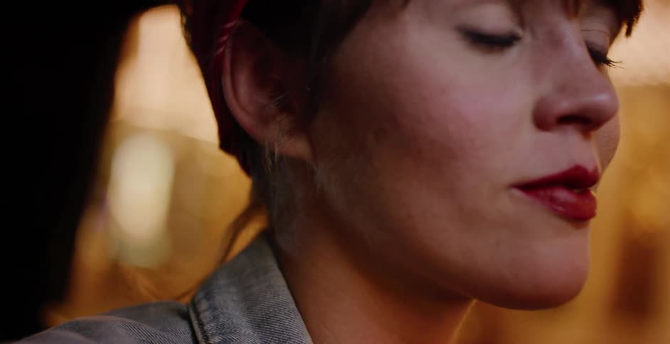A slow-motion close-up of a woman with red lipstick and a bandana looking thoughtfully off-camera in a warmly lit, intimate setting