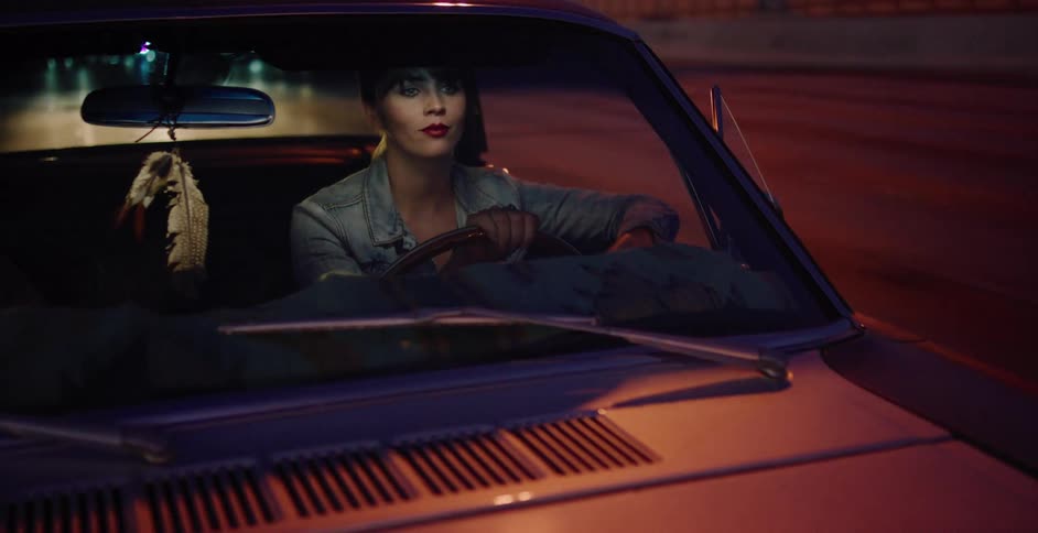 A stylish woman drives a vintage car on a city highway at night, with lights reflecting on the windshield