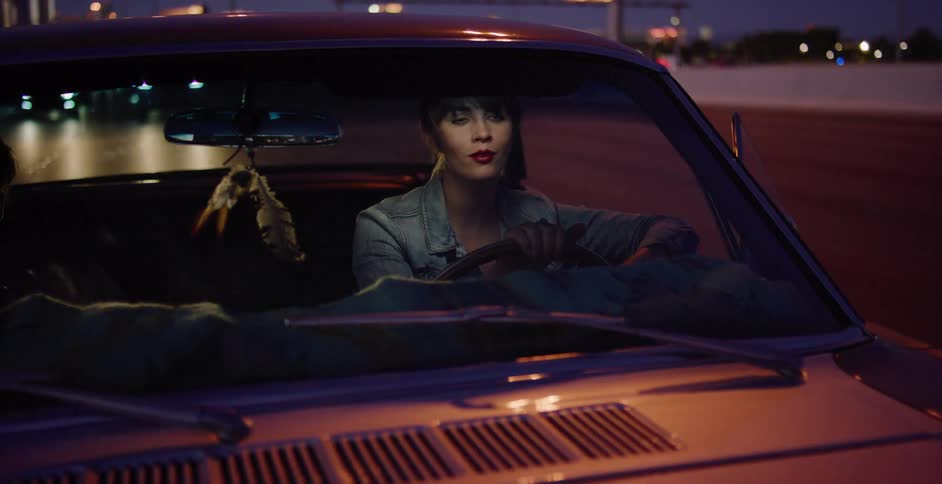 A cinematic shot of a stylish woman sitting in her classic car at night, looking around with city lights in the background