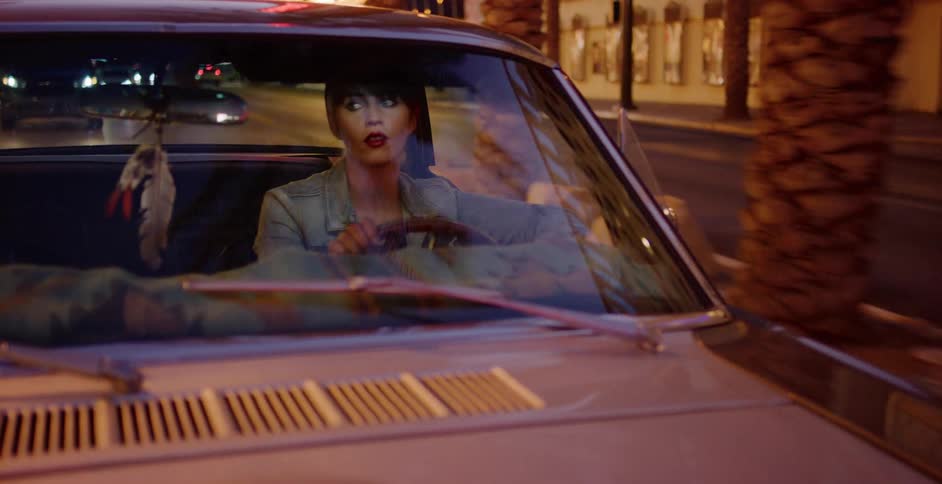 A stylish woman drives a classic car through the city at night, the streetlights reflecting in the windshield