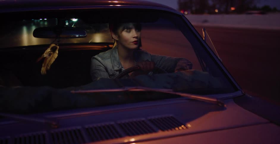 A stylish young woman sits behind the wheel of a classic car at night, bathed in the glow of passing city lights