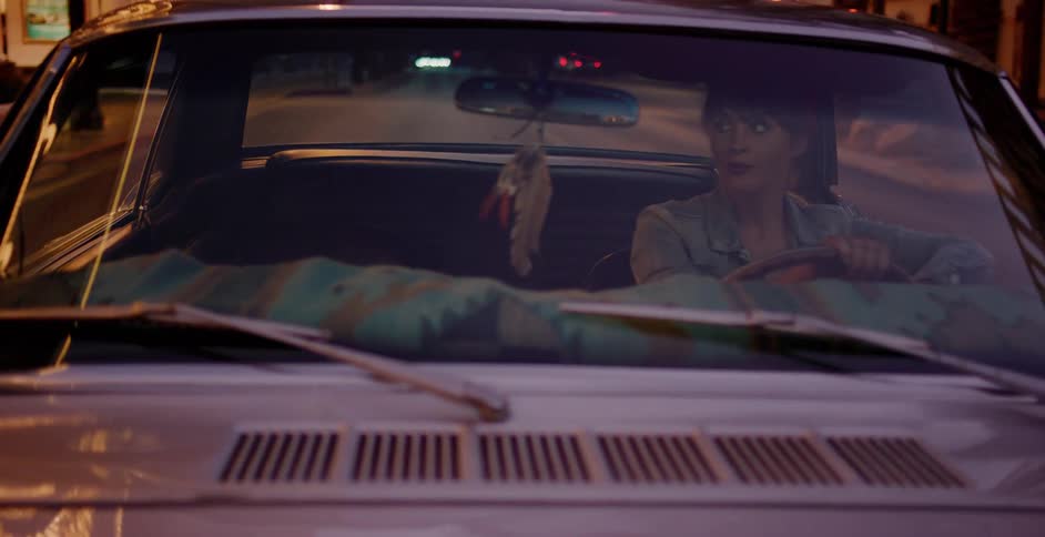 A cool young woman drives her vintage car through the city at night as the streetlights reflect off the windshield