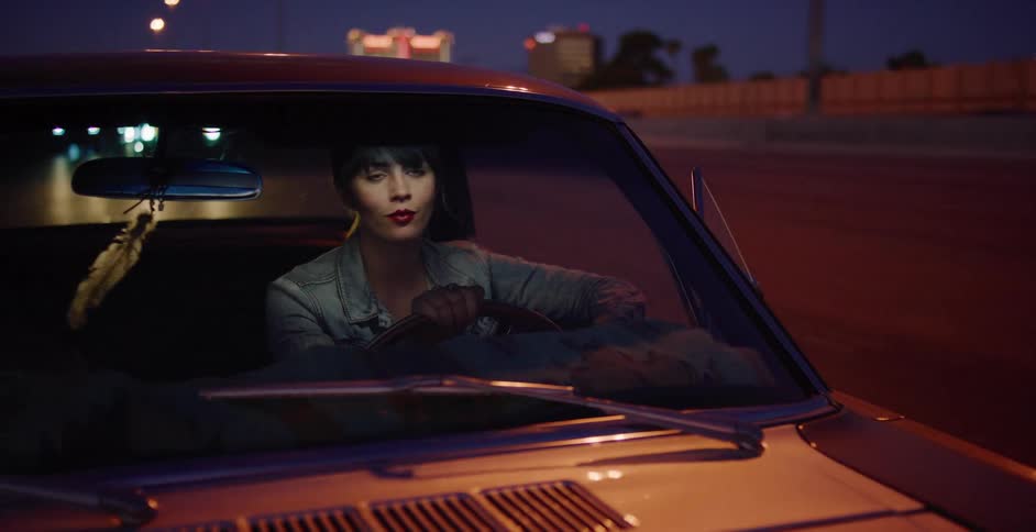 A cool woman with red lipstick drives a vintage car down a city freeway at dusk