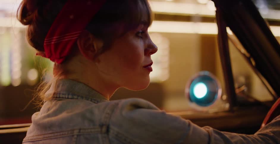A stylish woman in a denim jacket and red bandana drives her classic car through the bright city lights at night