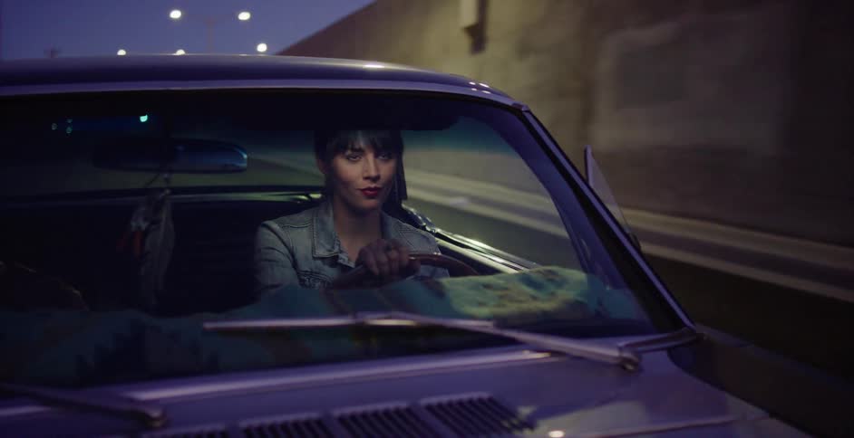 Front view of a stylish young woman driving her classic car down a city highway at night, her face illuminated by passing lights