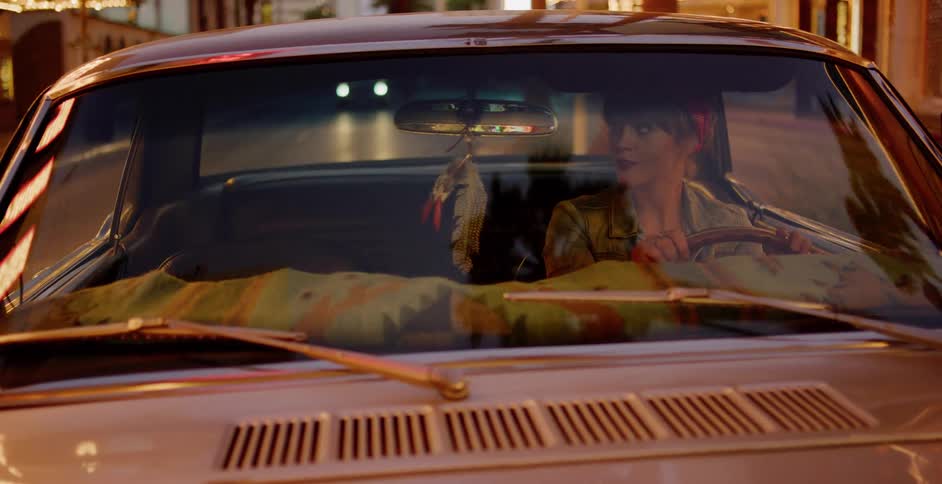 A stylish woman drives her vintage car through the city at night, with street lights reflecting on the windshield