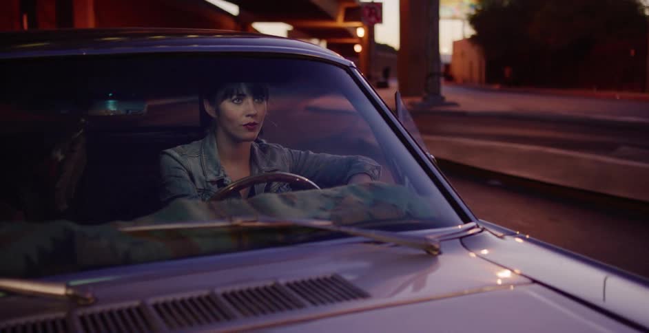 A stylish woman waits behind the wheel of her classic car on a city street as the evening lights reflect on the windshield