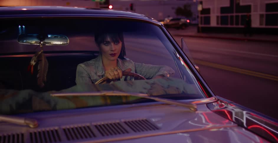 A stylish woman in a denim jacket sits behind the wheel of her vintage car on a city street at night, with pink and purple lights reflecting on the windshield