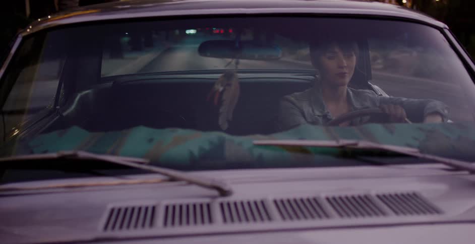 A young woman drives her classic car through the city at night, with streetlights and palm trees reflecting on the windshield