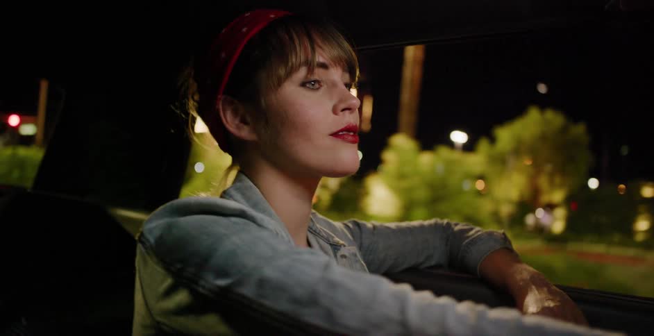 A stylish woman in a denim jacket and red bandana enjoys a late-night drive, watching the vibrant city lights pass by from the car