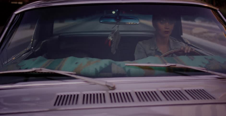 A woman drives her vintage muscle car through the city streets as evening sets in, seen through the windshield