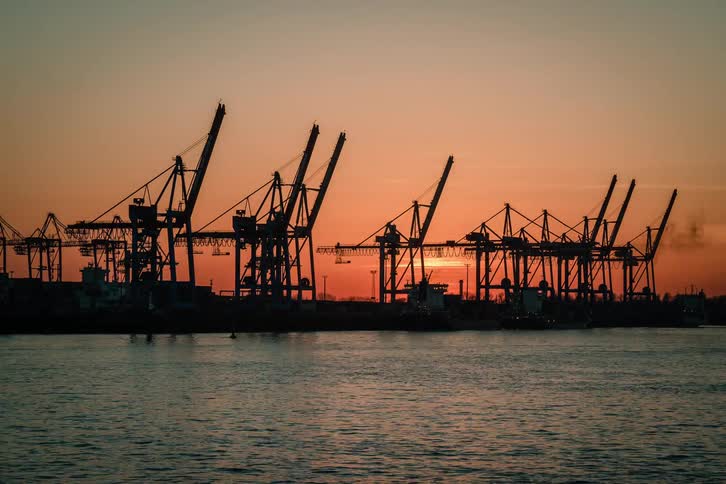 A captivating time-lapse captures the sun setting behind industrial cranes in a bustling harbor, silhouetting passing boats and ships against the vibrant twilight sky