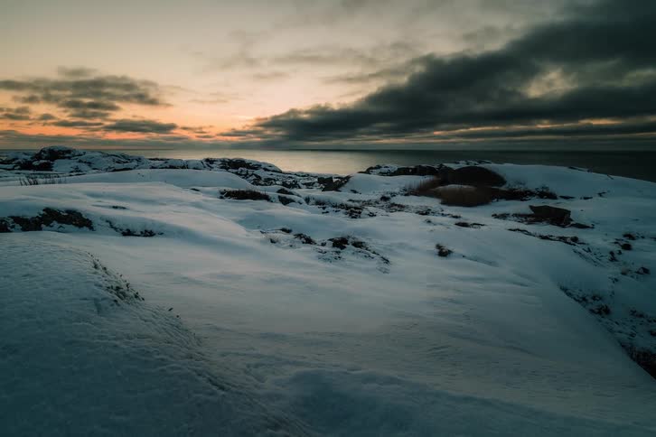A dramatic timelapse shows moody clouds moving over a snowy coastline as the sun sets over the ocean