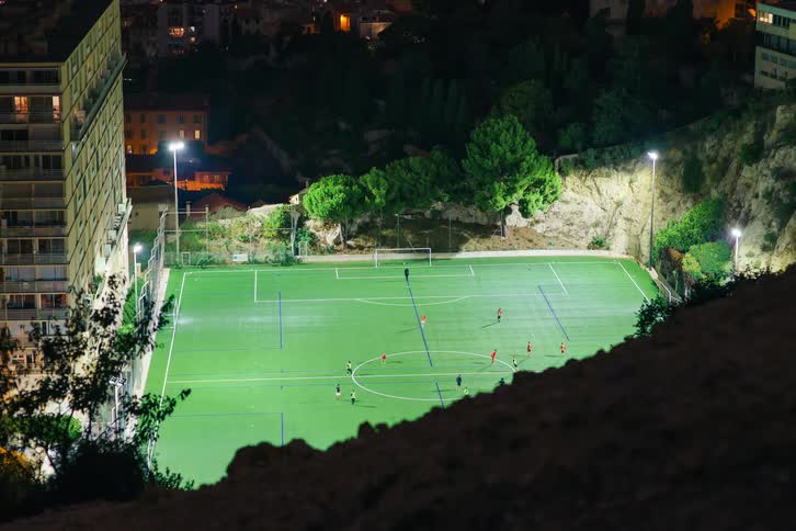 A high-angle timelapse captures a nighttime soccer game unfolding on a brightly lit field in the middle of a city
