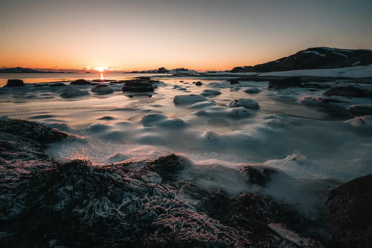The sun sets in a brilliant timelapse over a frozen arctic coastline, transitioning from golden hour to dusk