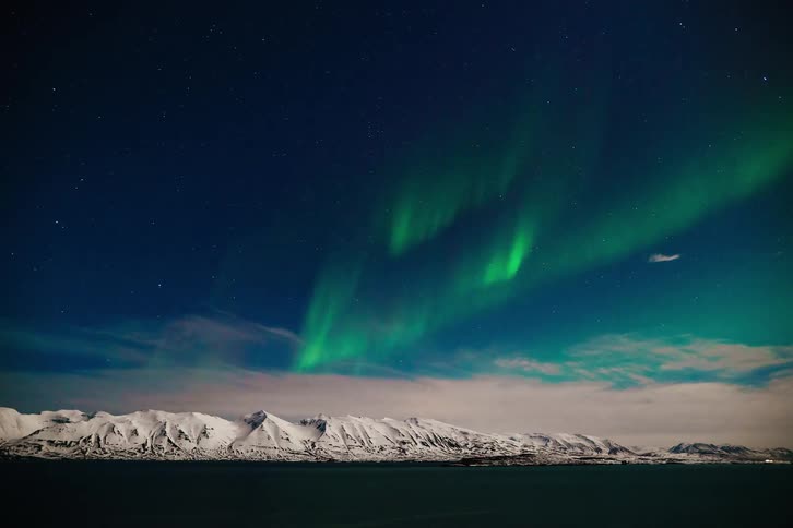 A stunning time-lapse captures the vibrant green Northern Lights dancing in the starry night sky over a snow-covered mountain range in Iceland