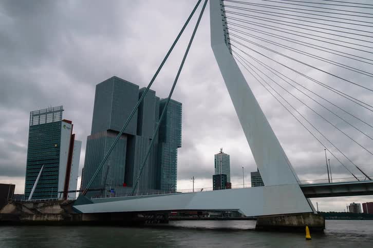 Time-lapse tilts up from the river to the Erasmus Bridge with the modern De Rotterdam building under a dramatic cloudy sky