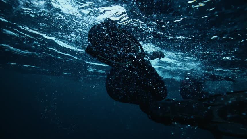 An ethereal slow-motion underwater view shows divers in wetsuits kicking their fins near the water's surface